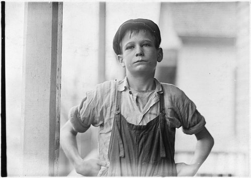 A young boy in work overalls stands with hands on hips, looking confidently at the camera.