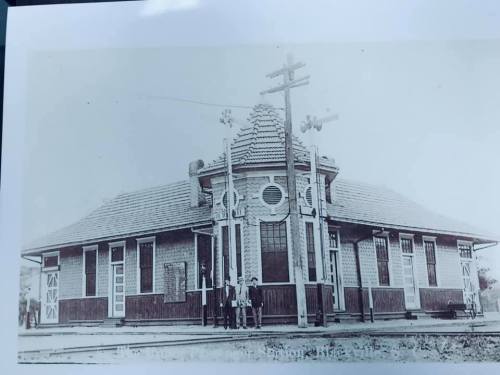 Historic black-and-white photo of a train station building with decorative architecture and two people standing outside.