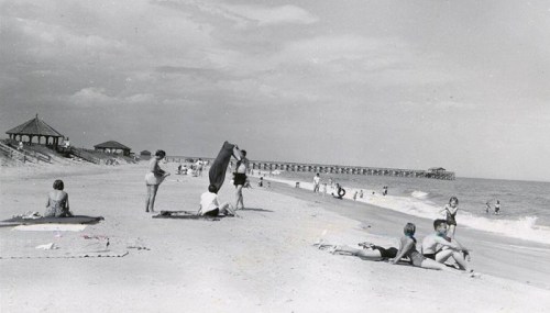 Black and white photo of a beach scene with people sunbathing, playing, and a pier in the background.