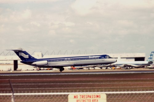 A blue and white airplane takes off from an airport runway, with hangars and a "No Trespassing" sign in the foreground.
