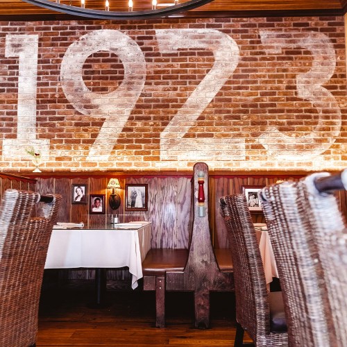 Interior of a restaurant featuring a brick wall with "1923" painted on it and woven chairs around wooden tables.