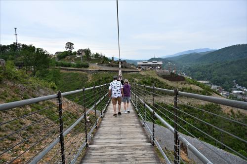 A couple walks on a wooden suspension bridge with scenic hills and buildings in the background.