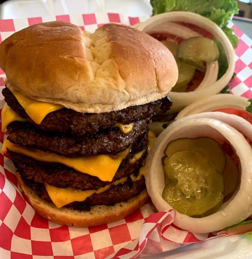 A towering cheeseburger with multiple beef patties and melted cheese, served with lettuce, tomato, and pickles on the side.