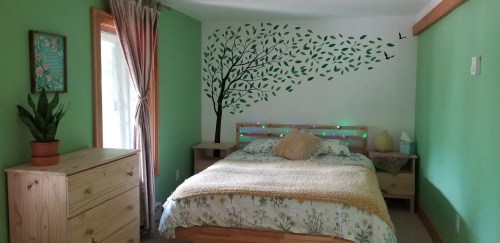 Cozy bedroom with green walls, a tree mural, a bed with decorative pillows, and a plant by the window.
