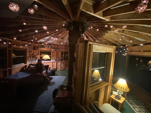 Cozy, circular wooden room with a bed, bookshelves, and warm string lights illuminating the space.