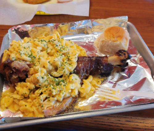 A tray with a smoked turkey leg topped with creamy macaroni and cheese, alongside a small dinner roll.