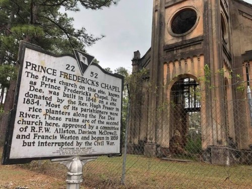 Historic sign for Prince Frederick's Chapel, with the chapel's ruins visible in the background, surrounded by trees.
