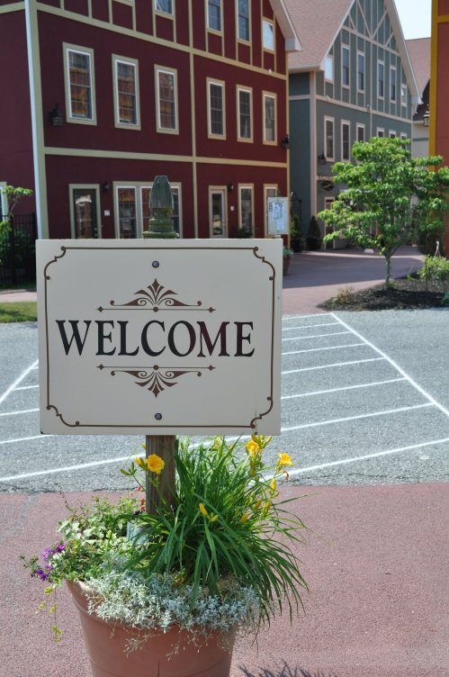 A "Welcome" sign stands in front of colorful buildings, surrounded by flowers in a pot.