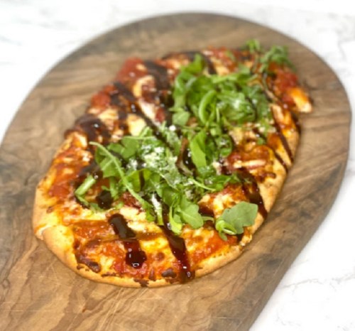 A flatbread pizza topped with sauce, cheese, arugula, and drizzled with balsamic glaze on a wooden serving board.