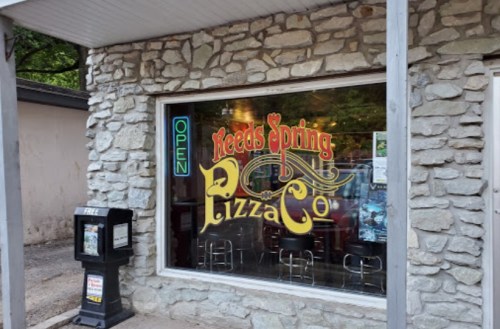 A stone building with a colorful "Open" sign and "Reeds Spring Pizza Co" in the window, featuring bar stools inside.