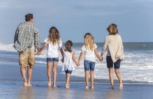 A family of five walks hand-in-hand along the beach, with waves gently lapping at their feet.