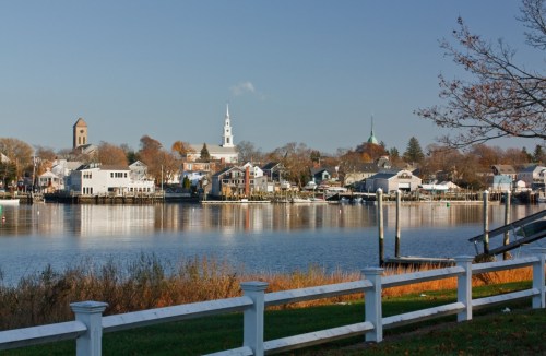 A serene waterfront view of a small town with churches, houses, and trees reflected in calm water under a clear sky.