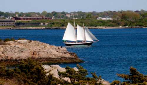 A sailboat with white sails glides through blue waters near rocky shores and green hills in the background.