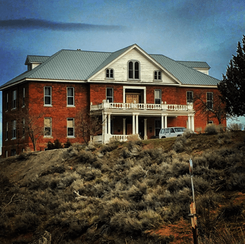 A large, two-story brick building with a gray metal roof, set on a hillside with sparse vegetation around it.