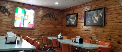 Cozy diner interior with wooden walls, colorful stained glass window, and orange chairs around a light blue table.