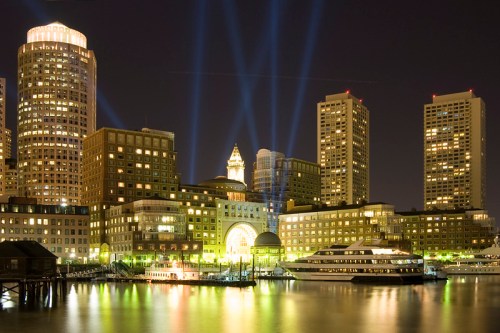 City skyline at night with illuminated buildings and beams of light reflecting on the water.