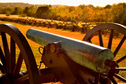 A close-up of a historic cannon in a field, with trees and a wooden fence in the background under a golden sunset.