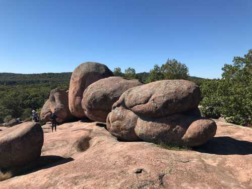 Two people stand near large, rounded boulders on a rocky surface, surrounded by trees and a clear blue sky.