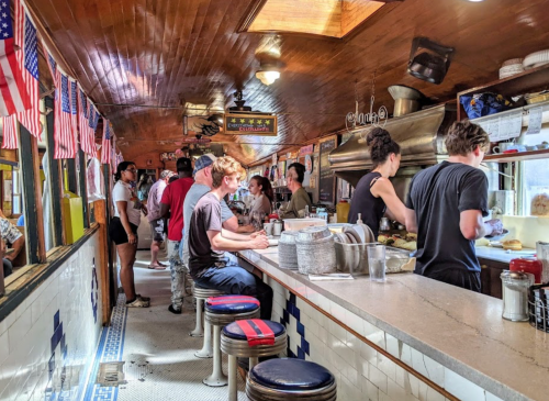 A bustling diner interior with patrons at the counter and staff preparing food, decorated with American flags.