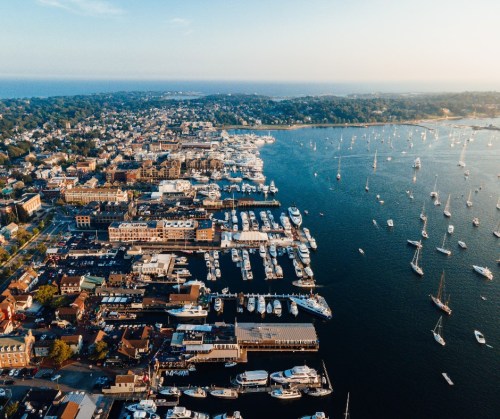 Aerial view of a bustling harbor with numerous boats, surrounded by a scenic coastal town and lush greenery.