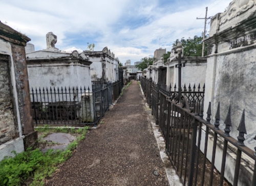 A narrow path between old, weathered tombs in a cemetery, surrounded by wrought iron fences and overgrown grass.