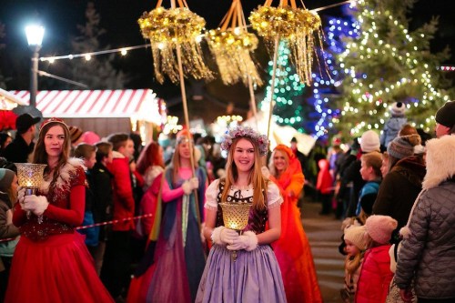 A festive crowd at night, with women in colorful dresses holding lanterns, surrounded by holiday lights and decorations.
