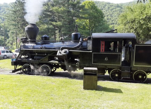 A vintage steam locomotive with smoke billowing, set against a backdrop of green trees and hills.