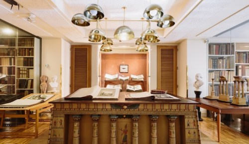 A cozy library with books, sculptures, and elegant lighting, featuring a large table with open books in the foreground.
