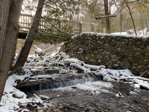 A serene winter scene featuring a small waterfall, snow-covered rocks, and a wooden bridge in a forested area.