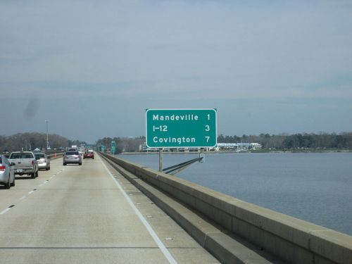 Highway sign indicating distances to Mandeville (1 mile), I-12 (3 miles), and Covington (7 miles) over a waterway.