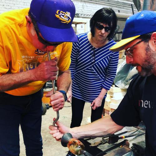 Two people work on glassblowing while a woman observes, all in a workshop setting. Tools and materials are visible.