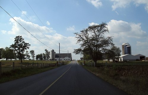 A rural road leads to a farm with a white barn, silos, and trees under a partly cloudy sky.