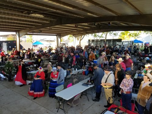 A large crowd of people in cowboy attire gathers under a covered area, with tables and tents in the background.