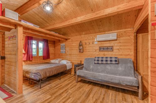 Cozy wooden cabin interior with a bed, futon, and rustic decor, featuring warm lighting and wooden walls.