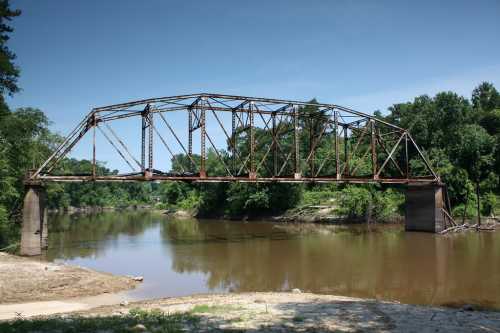 Rusty metal bridge arching over a calm river, surrounded by lush green trees under a clear blue sky.