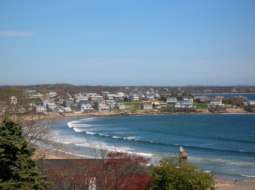 A coastal view featuring a beach, gentle waves, and a charming seaside town with houses and greenery in the background.