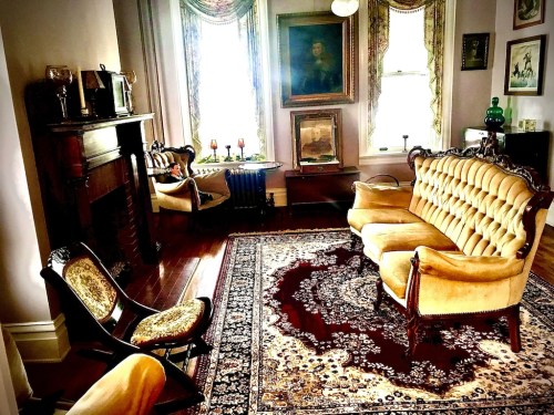 A vintage living room with a yellow tufted sofa, ornate chairs, a fireplace, and a decorative rug. Natural light filters in.