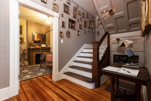 A cozy entryway with a staircase, framed photos on the wall, and a glimpse of a room beyond. Warm wooden floors.