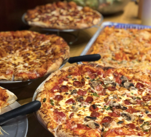 A variety of pizzas on display, featuring toppings like pepperoni, mushrooms, and cheese, on metal trays.