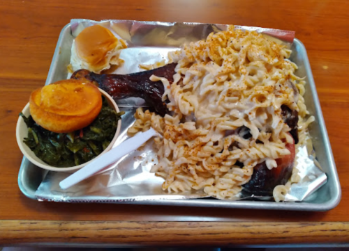 A tray with a large chicken leg, pasta topped with cheese, collard greens, and a small roll.