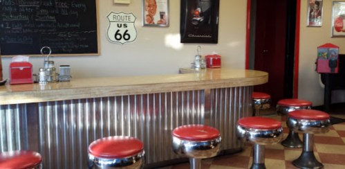 A retro diner interior featuring a chrome bar, red stools, and Route 66 signage on the wall.