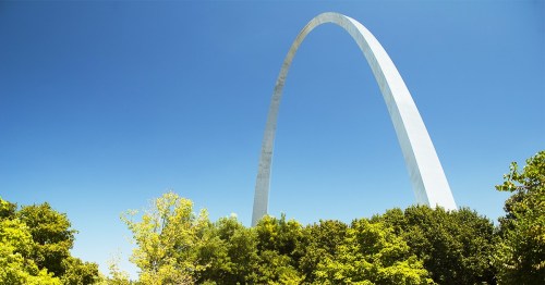 A tall, white arch structure rises against a clear blue sky, surrounded by green trees.