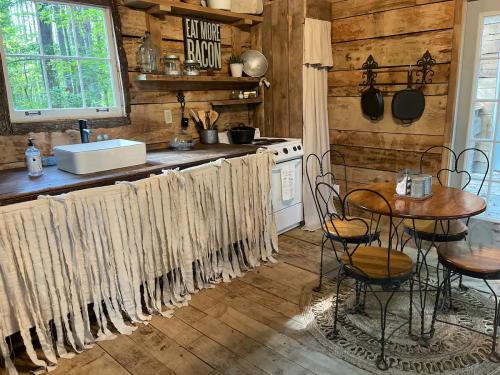 Cozy rustic kitchen with wooden walls, a white sink, and a round table with metal chairs, featuring a bacon-themed sign.