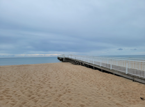 A sandy beach with a wooden pier extending into a calm, overcast sea.