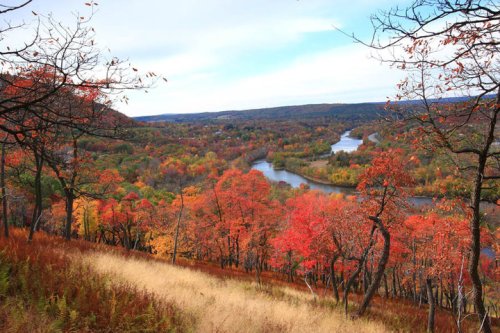 A scenic view of a river winding through vibrant autumn foliage and rolling hills under a cloudy sky.