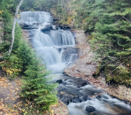 A serene waterfall cascades down rocky steps, surrounded by lush green trees and autumn leaves.