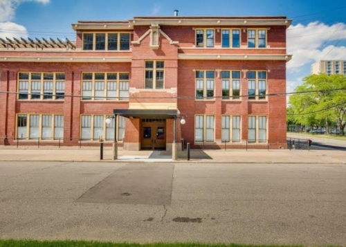 A red brick building with large windows, featuring a covered entrance and a grassy area in front.