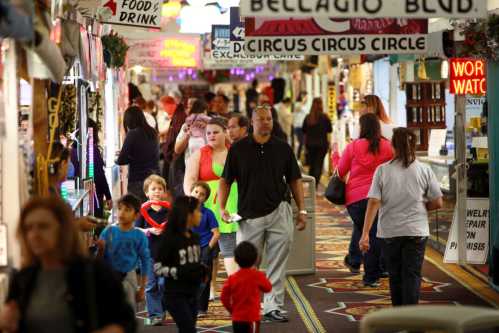 A busy indoor market with people walking, shops, and colorful signs, including families and children.