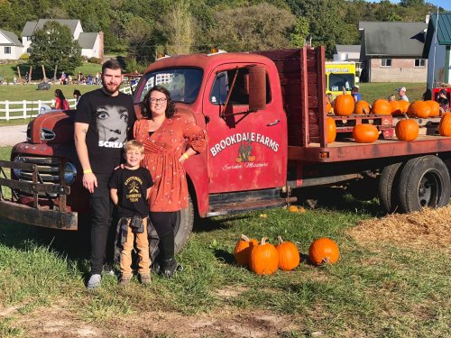 A family stands by a vintage red truck at a pumpkin patch, surrounded by pumpkins and a scenic farm backdrop.