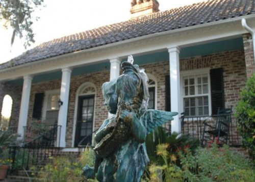 A vintage home with a brick facade and a mermaid statue in the foreground, surrounded by greenery.